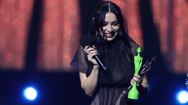 En leende Charli XCX håller i en mikrofon i en hand och två priser i en annan hand.