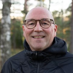 Kaljupäinen silmälasipäinen mies seisoo metsässä tummassa takissa, taustalla puita ja vaaleaa ruohoa.