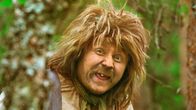 Trollet Rölli tittar fram bakom ett träd och grimaserar.