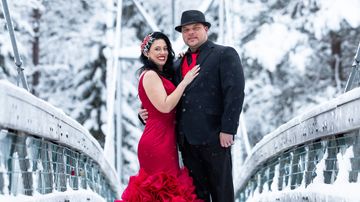 Punaiseen pukeutunut morsian ja sulhanen hymyilevät ja poseeraavat lumisella sillalla.