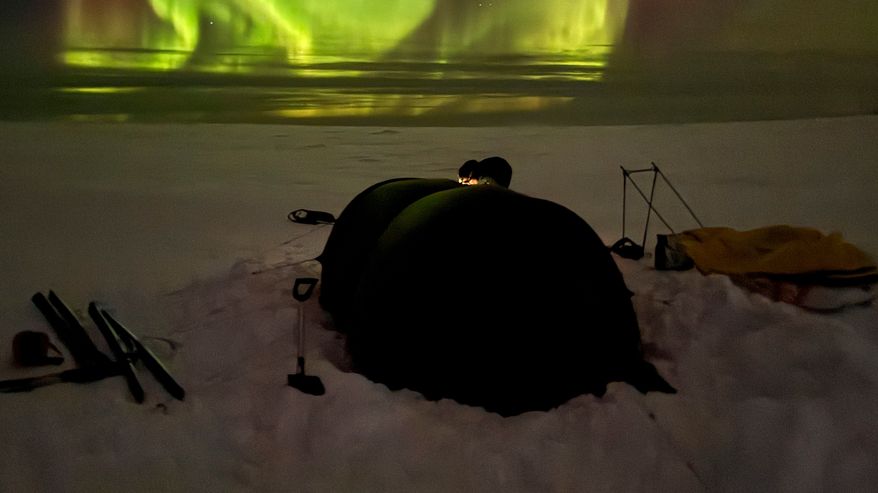 Revontulet loimuavat taivaalla talvisen maiseman yllä, etualalla näkyy teltta lumisella tundralla.