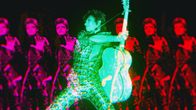 David Bowien vihreäksi sävytetty hahmo kitara kädessä, taustalla monistettuna punaisia Bowie.hahmoja; kuva elokuvasta Moonage Daydream.