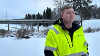 Mies seisoo ulkona kädet takin taskussa. Takki on keltainen ja siinä on heijastimia. Taustalla näkyy silta ja luminen maisema.