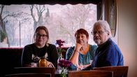 Kuvassa Hanna Eskelinen, Pauliina Rautio ja Jukka Korpela istuvat kahvilan pöydän ääressä. Kaikilla kolmella on keskittynyt ilme.