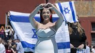 Ruskeahiuksinen nainen valkoisessa mekossa tekee käsillä sydämen ja pitää Israelin lippua.