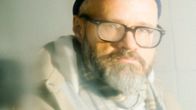 Miehellä on yllään ruskea takki, valkoinen paita, ruudullinen huivi, sekä sininen pipo ja silmälasit.