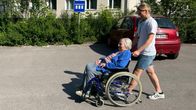Nuori tyttö työntää vanhaa rouvaa pyörätuolissa kerrostalon parkkipaikalla.