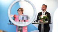 A woman in glasses and a pink blazer and a bearded man in a dark suit stand behind podiums with microphones, glasses of water and flowers.