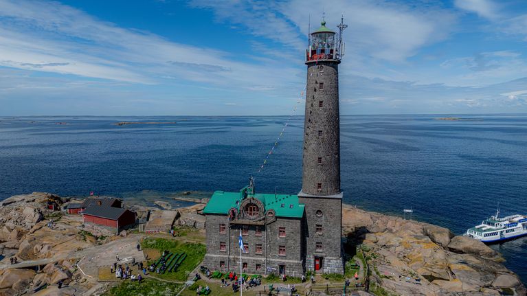 En fyr med tillhörande byggnad står på en klippig ö i havet, med en båt förtöjd vid bryggan och några människor på ön.