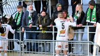 Två fotbollsspelare i vita dräkter, varav en med nummer 5 och namnet Kroviuson, står vid läktaren och möter supportrar i gröna halsdukar.