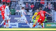 Fyra fotbollsspelare och en målvakt i gult samlas nära mål vid en match, med publiken synlig i bakgrunden.