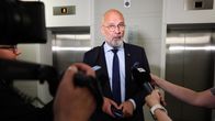 Bald, grey-bearded man in a dark suit, speaking to reporters.