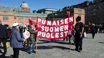 Vappumarssille osallistuvia ihmisiä Hakaniemen torilla. kaksi ihmistä pitelee banneria, jossa lukee "punaisen suomen puolesta".