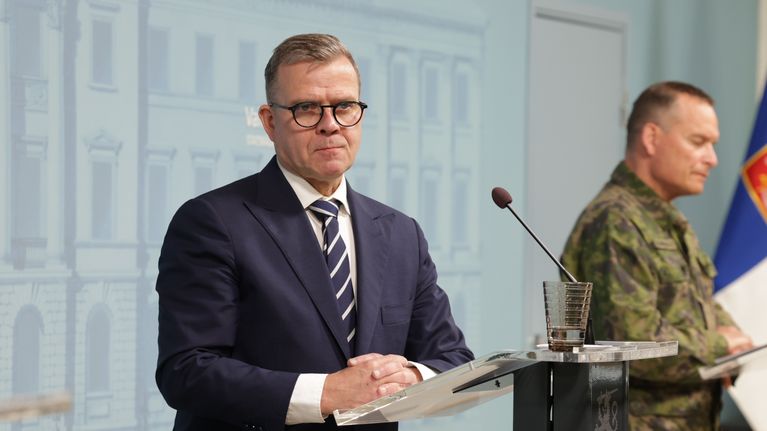 Petteri Orpo stands at a podium. A man dressed in camouflage stands in the background.