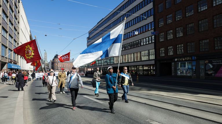 Vappumarssin osallistujat pitävät työväen lippuja ja Suomen lippua ylhäällä ja kävelevät kohti Pitkääsiltaa.