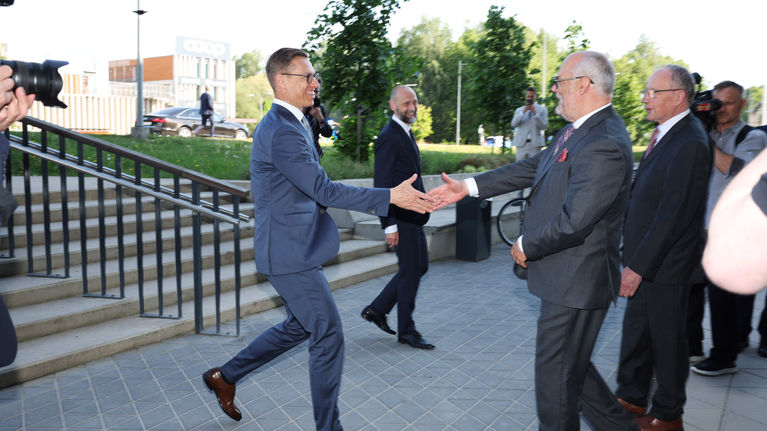 Two smiling men in dark suits shake hands outdoors in an urban area with trees and about 5 other men in the background.