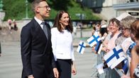 A man and a woman facing a group of people holding Finnish flags and smiling.