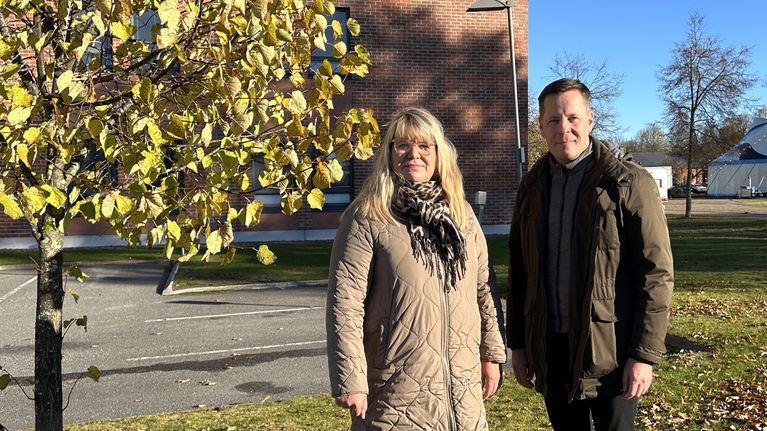 Kaksi henkilöä seisoo ulkona syksyisellä nurmikolla puun vieressä, taustalla tiilirakennus.