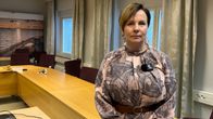 Nainen pukeutuneena kuvioituun mekkoon seisoo kokoushuoneessa, jossa taustalla näkyy pöytä ja tuoleja. Seinällä on maisemakuva.