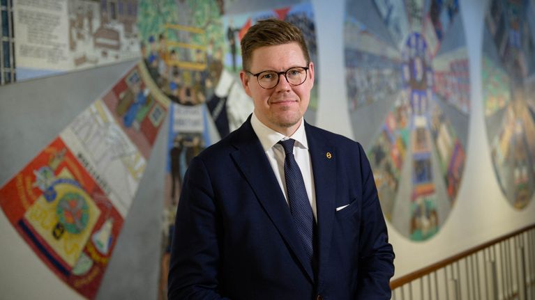Antti Lindtman i mörk kostym framför en mosaikvägg.