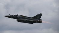 Sotilaslentokone Mirage 2000D lentää pilvisellä taivaalla, suihkumoottorin liekki näkyy koneen takana.