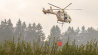 Pelastushelikopteri roikottaa vesitynnyriä lentäessään pensaiden ja puiden yläpuolella. 