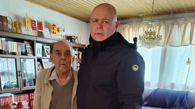 Två män, en äldre med käpp och en medelålders, står i ett vardagsrum med bokhylla, soffa och stolar.