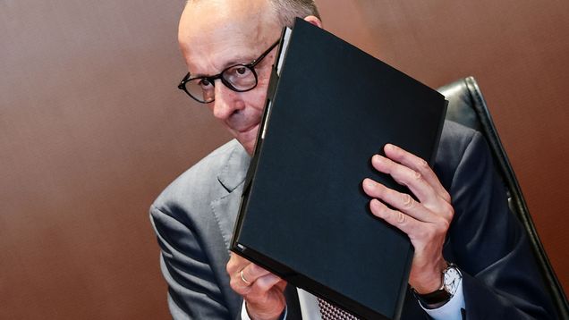 En man med glasögon i kostym håller en svart pärm framför sig, sittande mot en brun bakgrund.