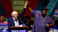 Senaattori Bernie Sanders ja rap-artisti Killer Mike seisovat puhujakorokkeella, hymyilevät ja pitävät toisiaan kädestä kiinni.