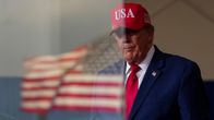 Donald Trump punaisessa "USA"-lippiksessä seisoo sisätiloissa; etualalla epätarkka Yhdysvaltain lippu.