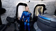 Astronautin pukuun pukeutunut vvaalea nainen istuu avaruusaluksessa ja katsoo hymyillen kameraan.
