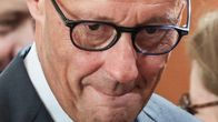 Mies pukeutuneena pukuun ja silmälaseihin katsoo alaviistoon. Taustalla epätarkkoja hahmoja.