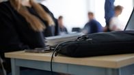 Puhelin ja koululaukku pöydällä. Taustalla tunnistamattomia oppilaita oppitunnilla. 