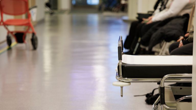Ihmisiä odottamassa pääsyä Kuopion yliopistollisen sairaalan päivystykseen. Etualalla sairaalasängyn pääty.