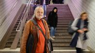 En person står i en tunnelbanestation iklädd en brun skinnjacka och en röd tröja. I bakgrunden syns människor som passerar på trappan.