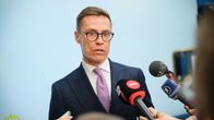 A man with brown hair and glasses, a dark jacket and lavender tie speaks into several handheld microphones. 