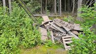 Kuva metsiköstä, jossa on maassa vanhoja puisia kuormalavoja ja muuta rakennusjätettä. Lavoista kasattua läjää ympäröi vihreää kasvillisuutta. Taustalla näkyy tiheä metsä, joka koostuu korkeista puista.