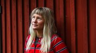 En person med ljust långt hår och röd randig tröja står framför en röd trävägg. 