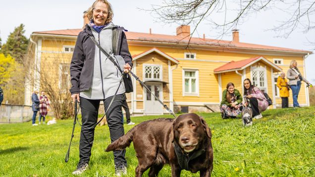 En person står på en gräsmatta med en hund i koppel framför ett gult trähus.