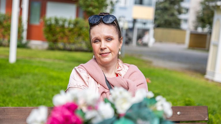 En kvinna sitter på en bänk utomhus med solglasögon på huvudet; blommor syns suddigt i förgrunden.