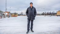 En person står på en isbana med flera bostadshus i bakgrunden. Det är vinter och marken runt omkring är täckt av snö.