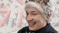 Samuli Souru nauraa. Hänellä on päässään permanenttirullat, jotka on kääritty muoviin. Kuvan taustalle on lisätty permanettirullia.