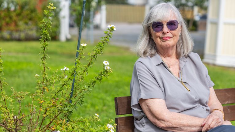 En äldre kvinna med grått hår och solglasögon sitter på en bänk i en trädgård, med grönska och blommor intill.
