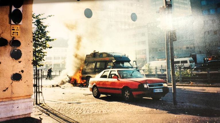 En röd Audibil står framför en brinnande bil på en stadsgata med rök och höga byggnader.