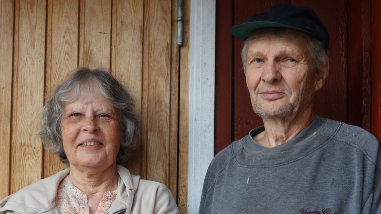 Sigbritt och Yngve Asplund, en kvinna och en man står bredvid varandra utomhus vid en röd trävägg och en dörr med glasruta.