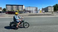 Mies ajaa vanhalla mopolla kadulla keltainen kypärä päässään, taustalla näkyy Kemin vanha kauppatori ja sen laitaan pystytettyjä vaalimainoksia.