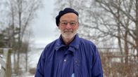 En man med skägg och glasögon iklädd en blå jacka står i ett snötäckt landskap.