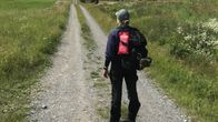 En person står på en väg med ryggen mot kameran. Personen har en keps på huvudet och en ryggsäck på ryggen där det hänger en brandfilt i sitt fodral. 