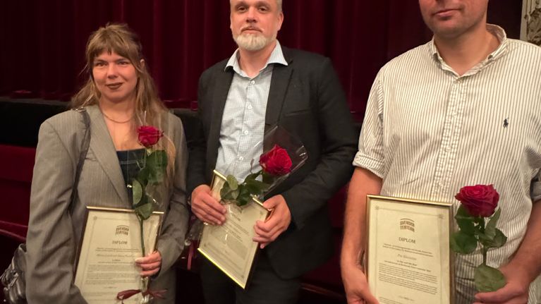 Tre prisbelönade personer med ros och diplom står på rad.