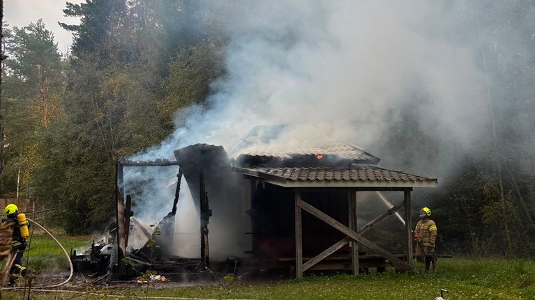 En byggnad i trä står i brand med kraftig rökutveckling, två brandmän arbetar på platsen.